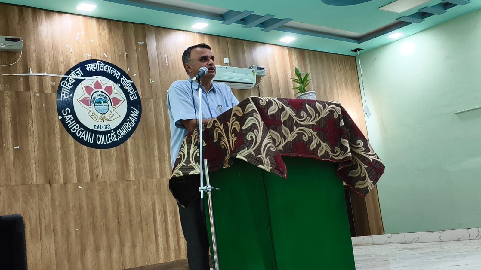 A speaker at a podium in front of the Sahibganj College seal