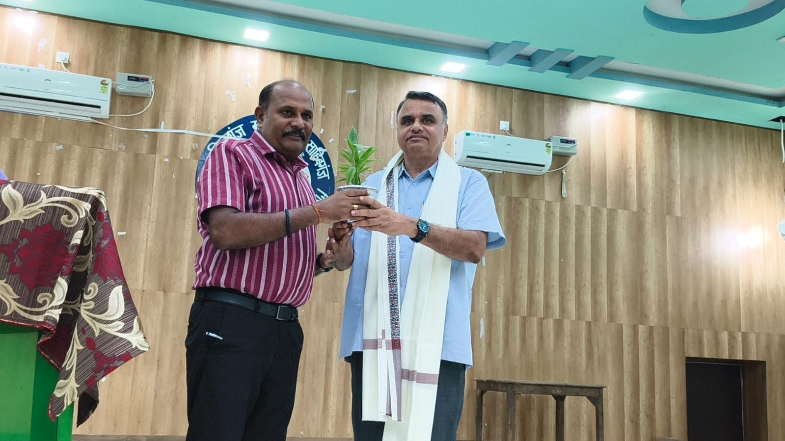 Two men on stage, one handing over a small plant, with the college seal on the wall